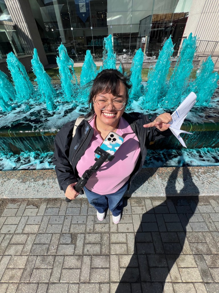 Business Support Specialist holding up a peace sign with tripod in hand and blue fountains in the background.