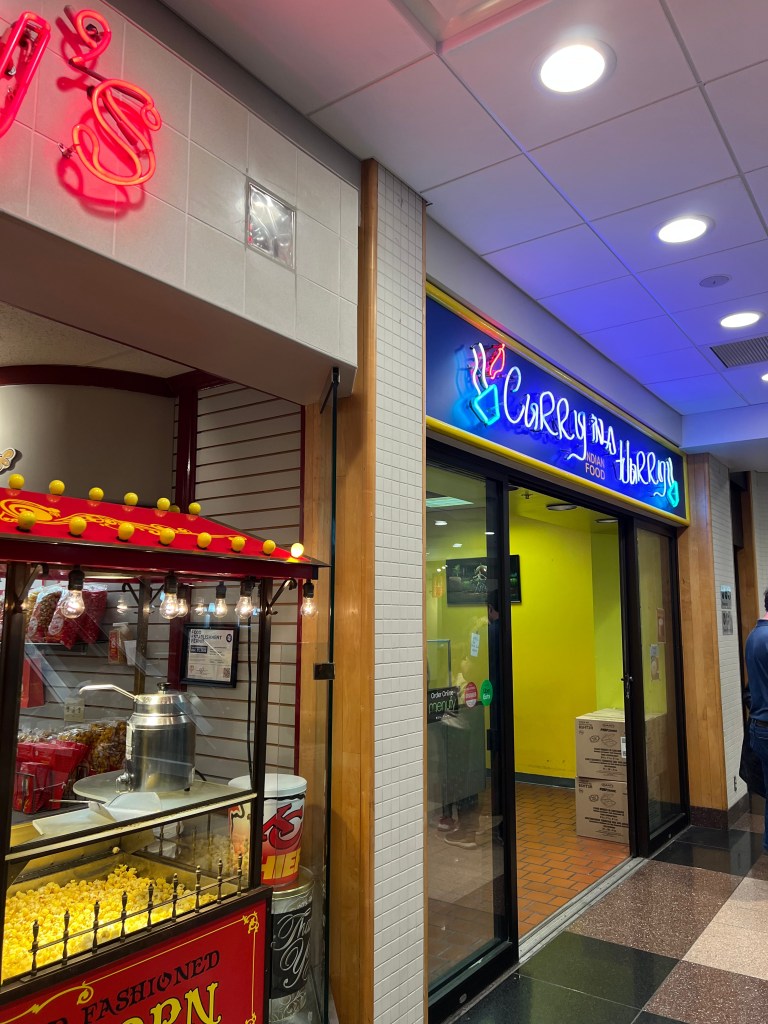 Neon signage for Topsy's Popcorn next to Curry in a Hurry neon signage, with popcorn cart.